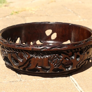 Puede incluir: Un cuenco de madera marrón oscuro con intrincados tallados de animales, posiblemente leones, alrededor del borde. El cuenco tiene un acabado liso y pulido y es perfecto para servir fruta o aperitivos.