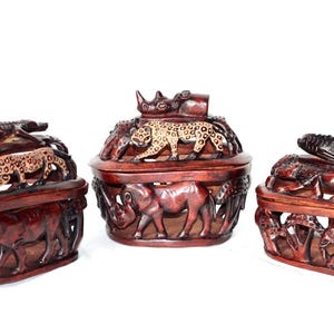 Könnte beinhalten: Drei Holzkisten mit aufwendigen Schnitzereien von afrikanischen Tieren. Die Kisten sind oval geformt und haben eine dunkel rotbraune Oberfläche. Zu den Tieren gehören ein Nashorn, ein Leopard, ein Krokodil, ein Elefant und eine Giraffe.