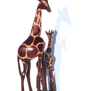 Op de afbeelding: Houten girafsculptuur met een moeder en een babygiraf. De giraffen zijn gesneden uit donkerbruin hout met lichtere bruine patroonmarkeringen. De sculptuur is gemonteerd op een donkere, rechthoekige basis, die Afrikaanse dieren in het wild vertegenwoordigt.