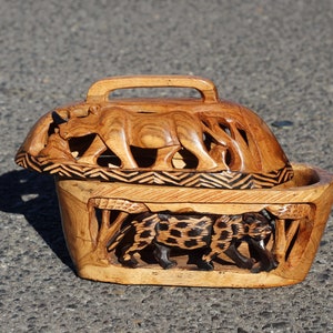 Puede incluir: Una caja de madera con una tapa tallada que representa un rinoceronte y un leopardo. La caja está hecha de madera marrón claro y tiene una mancha marrón oscura. La tapa tiene un asa y está decorada con un patrón geométrico.