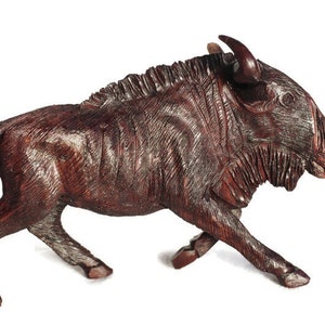 Può includere: Una scultura in legno intagliato di uno gnu in posa di corsa. La scultura è di un ricco colore marrone scuro con venature del legno visibili e intaglio dettagliato. Lo gnu ha corna e una criniera strutturata.