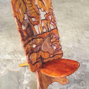 May include: Hand-carved wooden chair with a detailed relief of African wildlife, including elephants and a lion. The chair has a curved back and a small seat, all in warm brown tones. A unique piece of furniture.