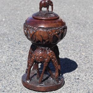 Puede incluir: Una caja de madera marrón oscuro con un elefante en la parte superior y tres jirafas en la base. La caja está tallada con intrincados detalles de animales africanos.