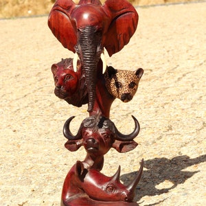 Op de afbeelding: Een houten sculptuur van een stapel Afrikaanse dieren. De sculptuur is gemaakt van donker rood hout en toont een olifant, een leeuw, een luipaard, een buffel en een neushoorn. De dieren zijn realistisch gebeeldhouwd en zijn in een gestapelde formatie gerangschikt.