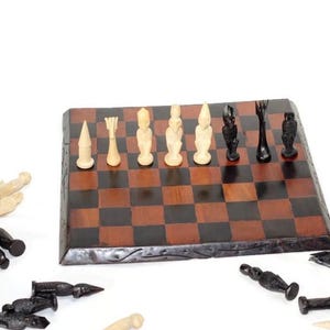 Può includere: Una scacchiera di legno con caselle nere e marroni. Pezzi degli scacchi bianchi e neri sono disposti sulla scacchiera. Altri pezzi sono sparsi intorno alla scacchiera.