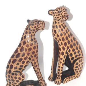 Op de afbeelding: Twee houten sculpturen van cheeta's met een gevlekt patroon in bruin- en beigetinten. De cheeta's zitten, met hun hoofden opzij gedraaid. De sculpturen staan op een witte achtergrond.