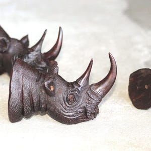 Può includere: Primo piano di sculture di teste di rinoceronte in legno marrone scuro con intagli dettagliati. Le teste di rinoceronte hanno corna prominenti e sono disposte su una superficie chiara. Si tratta probabilmente di oggetti decorativi o da collezione.