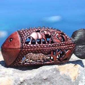 Puede incluir: Una caja de madera tallada con forma de pez con detalles intrincados. La caja de color marrón rojizo presenta diseños recortados de animales africanos, incluyendo elefantes, un leopardo y un búfalo. La caja descansa sobre una roca gris con un fondo azul oceánico.