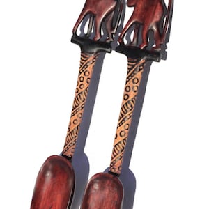 Op de afbeelding: Twee houten opscheplepels met gesneden dierenfiguren op de handvatten. De lepels zijn donkerrood met een decoratief patroon op de handvatten. Eén lepel is een vork, de andere is een lepel.