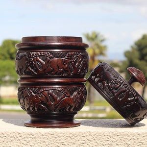 Puede incluir: Un recipiente de madera marrón oscuro con tapa, tallado intrincadamente con un patrón repetido de animales. El recipiente es cilíndrico y tiene una base redondeada. La tapa tiene un pomo en la parte superior.