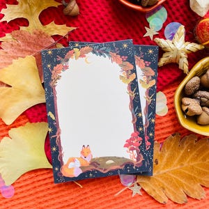 Op de afbeelding: Twee briefkaarten met een bos-thema rand met een vos illustratie. De kaarten liggen op een oranje oppervlak met herfstbladeren, eikels en decoratieve kommen. De rand heeft een donkerblauwe achtergrond met sterren.