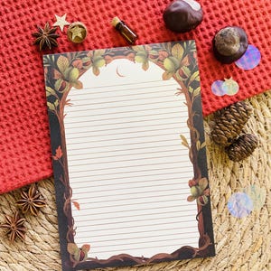 May include: Lined writing paper with a decorative border of trees and foliage. The paper is white with horizontal lines. The border is brown, green, and gold. The stationery is on a red textured surface.