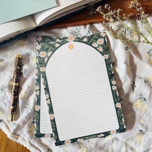 Peut inclure: Deux blocs-notes lignés avec un motif de marguerite et de soleil. Les blocs-notes ont une bordure vert foncé avec des marguerites blanches et un soleil en haut. Un stylo à motifs et un livre ouvert sont également dans l'image, avec une brindille de fleurs séchées.