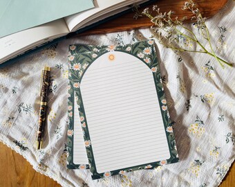 Puede incluir: Dos blocs de notas rayados con un diseño de margarita y sol. Los blocs tienen un borde verde oscuro con margaritas blancas y un sol en la parte superior. Un bolígrafo estampado y un libro abierto también están en la imagen, junto con una ramita de flores secas.