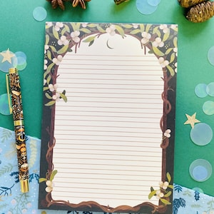 May include: Lined notepad with a decorative tree and floral design. The notepad features a brown border with a tree-like archway, white berries, and a crescent moon. A pen with a celestial design is on the left. The background is green.