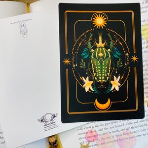 May include: A black and gold illustrated card features a green and gold beetle with a crown on its head. The beetle is surrounded by a circle of stars and a crescent moon. The card has a gold border and a white background.
