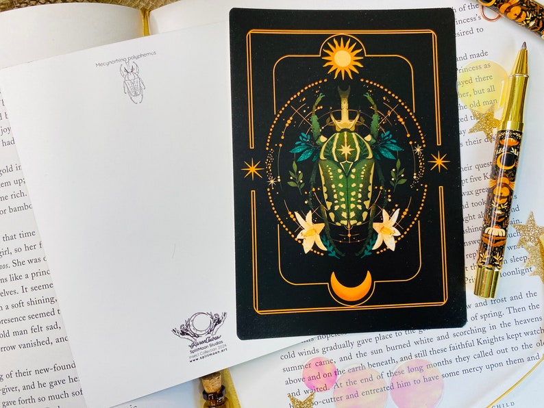 May include: A black and gold illustrated card features a green and gold beetle with a crown on its head. The beetle is surrounded by a circle of stars and a crescent moon. The card has a gold border and a white background.
