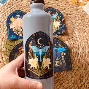 K&ouml;nnte beinhalten: Eine graue wiederverwendbare Wasserflasche mit schwarzem Deckel und Griff. Die Flasche hat einen Aufkleber mit einem blauen Schmetterling, wei&szlig;en Blumen und einem Halbmond auf schwarzem Hintergrund.