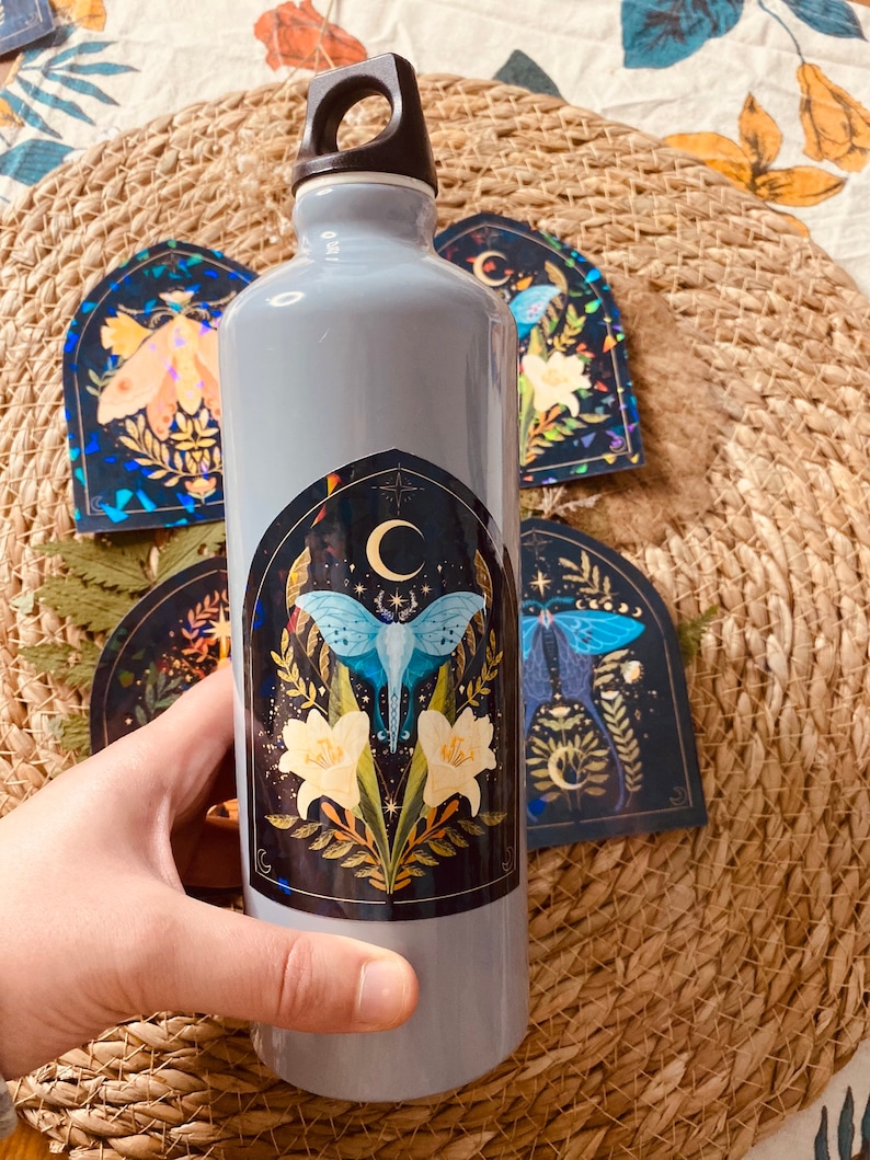 K&ouml;nnte beinhalten: Eine graue wiederverwendbare Wasserflasche mit schwarzem Deckel und Griff. Die Flasche hat einen Aufkleber mit einem blauen Schmetterling, wei&szlig;en Blumen und einem Halbmond auf schwarzem Hintergrund.