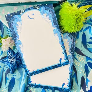 May include: Two blank notepads with a blue floral design and a crescent moon and star. The notepads are on a blue patterned fabric with a green and yellow flower and a blue star-shaped decoration. The notepads have a scalloped edge.