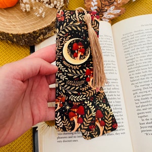 May include: A black and gold bookmark with a crescent moon and red mushrooms on a dark background. The bookmark has a tassel.