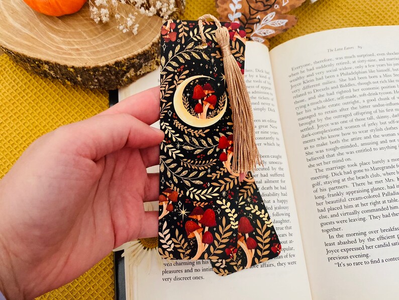 May include: A black and gold bookmark with a crescent moon and red mushrooms on a dark background. The bookmark has a tassel.