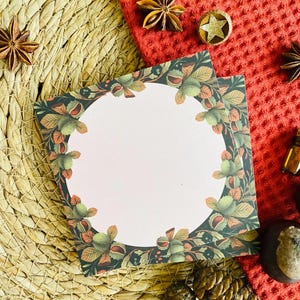 May include: Two square cards with a floral design featuring a light pink center and a border of green leaves and nuts. The cards are on a woven mat with star anise, a red cloth, and decorative elements.
