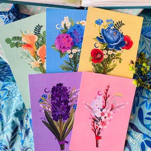 Peut inclure: Plusieurs impressions d'art floral avec des bouquets colorés sur des fonds pastel. Les impressions présentent des illustrations détaillées de fleurs dans des tons orange, bleu, violet et rose. L'œuvre est disposée sur un tissu à motifs floraux bleus.
