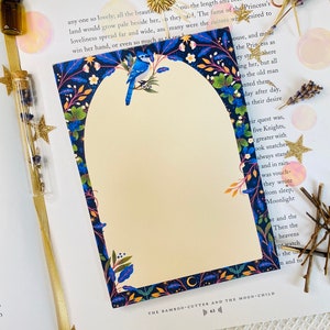 Puede incluir: Bloc de notas decorativo con una ilustración de un pájaro azul y un borde floral en azul, verde y blanco. El bloc de notas está sobre un libro abierto, con un marcapáginas dorado y decoraciones de estrellas.