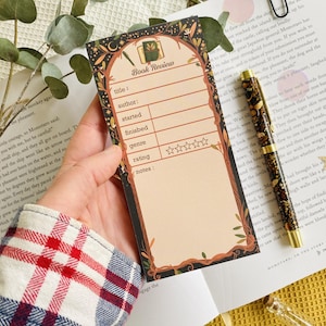 May include: A decorative book review notepad with a floral and botanical design. The notepad has spaces for title, author, start and finish dates, genre, rating, and notes. A pen and book are also in the image.