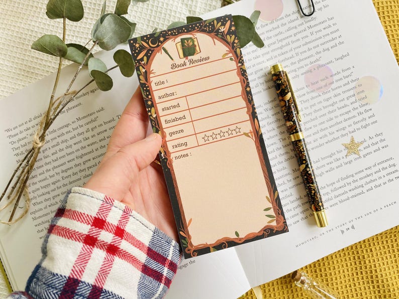 May include: A decorative book review notepad with a floral and botanical design. The notepad has spaces for title, author, start and finish dates, genre, rating, and notes. A pen and book are also in the image.