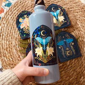 K&ouml;nnte beinhalten: Eine graue wiederverwendbare Wasserflasche mit einem Aufkleber eines blauen Nachtfalters mit gelben Blumen und einem Halbmond auf einem dunklen Hintergrund. Der Aufkleber hat ein gotisches Bogen-Design.