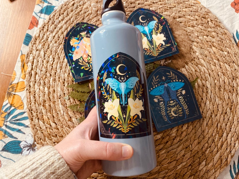 K&ouml;nnte beinhalten: Eine graue wiederverwendbare Wasserflasche mit einem Aufkleber eines blauen Nachtfalters mit gelben Blumen und einem Halbmond auf einem dunklen Hintergrund. Der Aufkleber hat ein gotisches Bogen-Design.