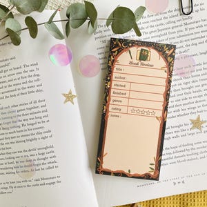 May include: A decorative bookmark with a black border and a beige interior, featuring the words "Book Review" and spaces to fill in details like title, author, and rating. The bookmark is set on an open book with a yellow surface.