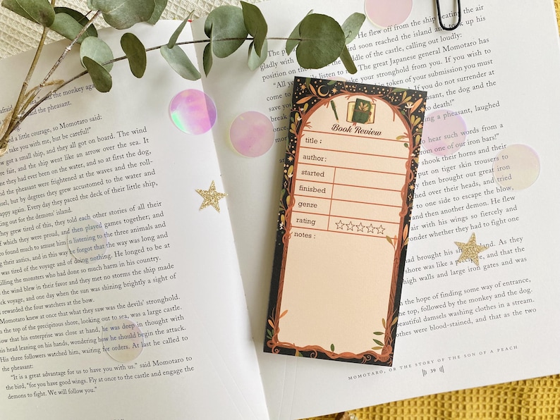 May include: A decorative bookmark with a black border and a beige interior, featuring the words "Book Review" and spaces to fill in details like title, author, and rating. The bookmark is set on an open book with a yellow surface.