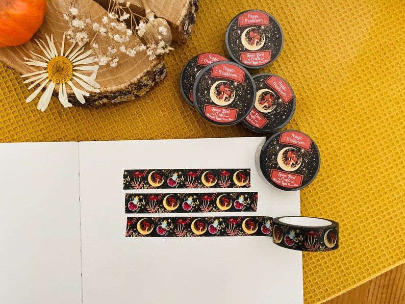 Op de afbeelding: Drie rollen zwarte en gouden washi tape met een herhalend patroon van rode paddenstoelen, halve manen en sterren. De tape is gelabeld met "Magic Mushrooms" en "Space Bird Crafts".