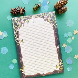 May include: A lined notepad with a decorative border featuring a tree branch design with white flowers and a crescent moon. The notepad is surrounded by decorative elements, including star anise, pine cones, and iridescent confetti, on a green surface.