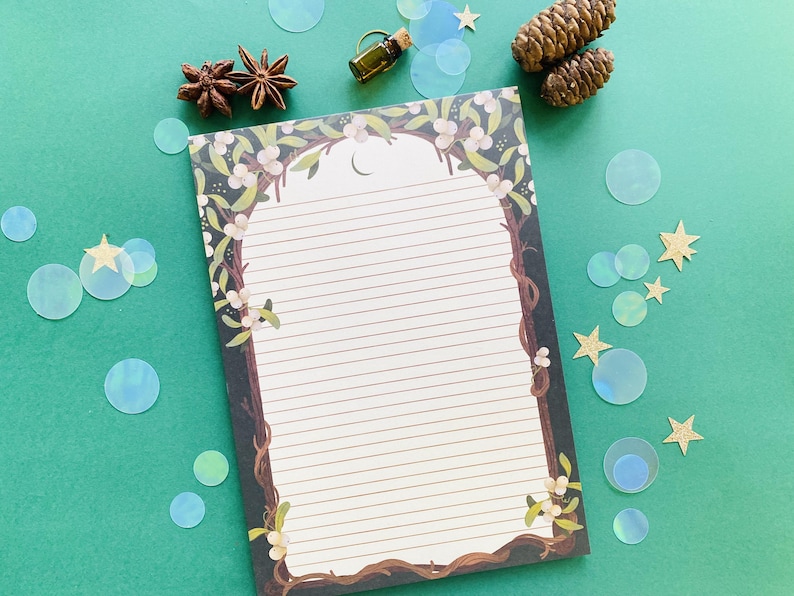 May include: A lined notepad with a decorative border featuring a tree branch design with white flowers and a crescent moon. The notepad is surrounded by decorative elements, including star anise, pine cones, and iridescent confetti, on a green surface.