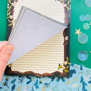 May include: A notepad with a decorative border featuring a brown tree branch design with white flowers. The notepad has lined pages and is being flipped open. The background includes a green surface with star and circle decorations.