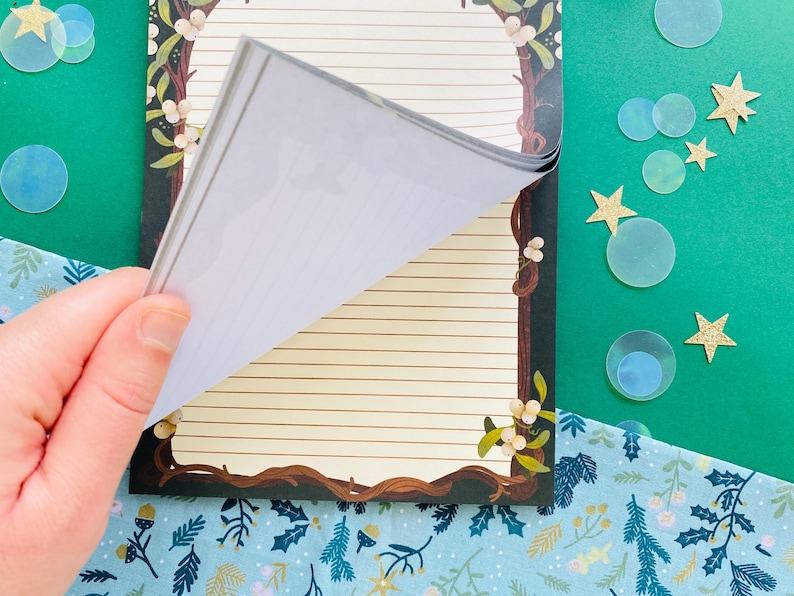 May include: A notepad with a decorative border featuring a brown tree branch design with white flowers. The notepad has lined pages and is being flipped open. The background includes a green surface with star and circle decorations.