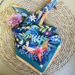 Puede incluir: Un pañuelo cuadrado de seda con un fondo verde azulado oscuro y un diseño floral y de polilla. El pañuelo tiene un borde naranja claro y está doblado sobre un salvamanteles tejido. Una flor rosa está colocada en el pañuelo.