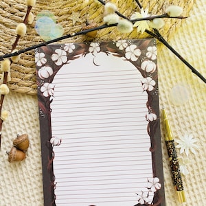 Könnte beinhalten: Liniertes Briefpapier mit einem Blumendesign mit weißen Baumwollblüten und braunen Ranken. Ein goldener und schwarzer Stift liegt neben dem Briefpapier. Das Briefpapier liegt auf einer strukturierten, cremefarbenen Oberfläche mit dekorativen Zweigen und Eicheln.