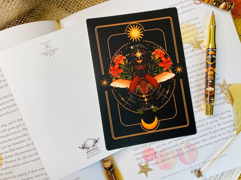 May include: A black and gold tarot card with a detailed illustration of a beetle with wings spread, surrounded by flowers and a celestial design. The card features a sun and moon in the corners.