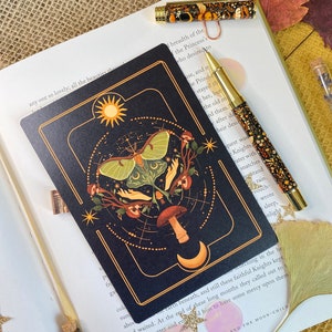 Peut inclure: Une carte noire avec une bordure dor&eacute;e repr&eacute;sente une lune, des &eacute;toiles, un champignon et un papillon de nuit vert aux ailes d&eacute;ploy&eacute;es. La carte porte l'inscription "The Bamboo-Cutter and the Moon-Child 63 III" en bas.