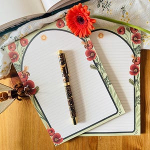 Può includere: Due blocchi per appunti a righe con un bordo floreale con papaveri rossi e api. Una penna decorativa con dettagli dorati poggia sul blocco per appunti. Sono presenti anche un'ape di legno e una margherita rossa. Gli oggetti sono disposti su una superficie di legno.