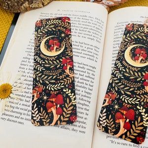 May include: A black bookmark with a gold crescent moon and mushrooms. The background is black with gold and white floral designs. The bookmark has a gold tassel.