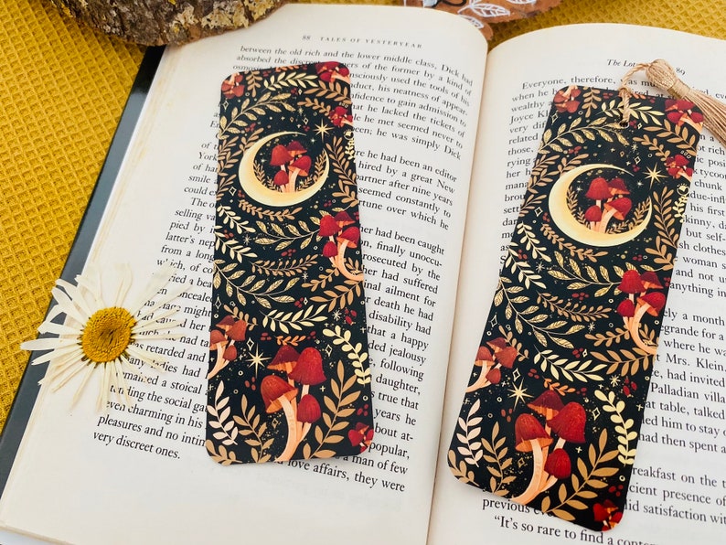 May include: A black bookmark with a gold crescent moon and mushrooms. The background is black with gold and white floral designs. The bookmark has a gold tassel.
