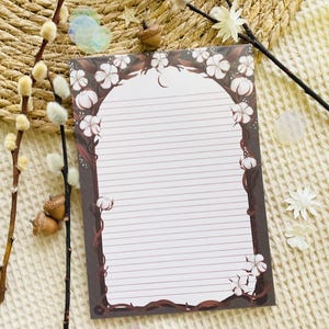 Pu&ograve; includere: Carta da lettere a righe con bordo floreale e di vite. Il design presenta fiori bianchi e batuffoli di cotone su sfondo marrone scuro. La carta &egrave; bianca con linee orizzontali per la scrittura. Elementi decorativi: ramoscelli e ghiande.