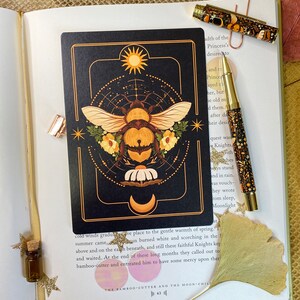 Peut inclure: Une carte noire avec une illustration dor&eacute;e et blanche d'une abeille avec un soleil et une lune en arri&egrave;re-plan. Le texte "The Bamboo-Cutter and the Moon-Child" est en bas de la carte.