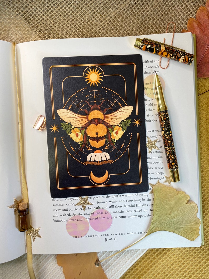Peut inclure: Une carte noire avec une illustration dor&eacute;e et blanche d'une abeille avec un soleil et une lune en arri&egrave;re-plan. Le texte "The Bamboo-Cutter and the Moon-Child" est en bas de la carte.